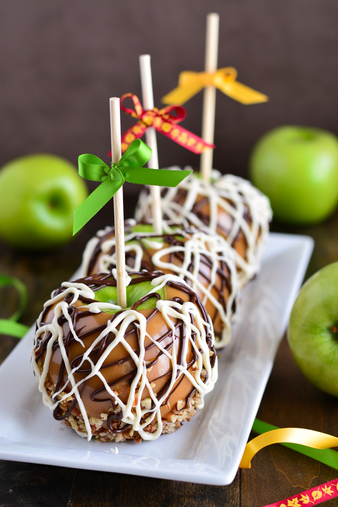 Three Caramel Apples decorated with drizzled chocolate, dipped in nuts, and ribbon bows on the sticks.
