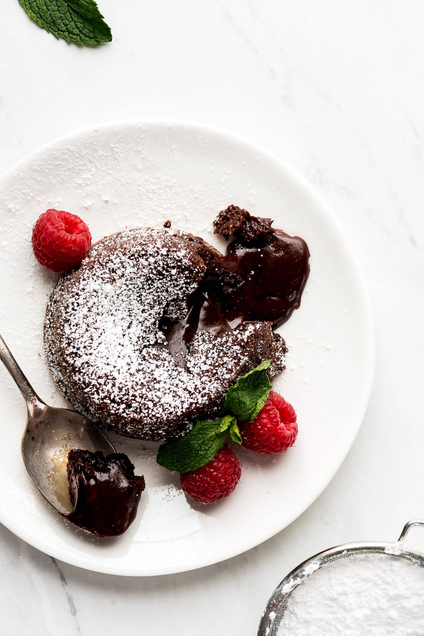 Chocolate Lava Cake on a plate with raspberries and a spoonful of the cake on a spoon with the lava flowing out onto the plate.