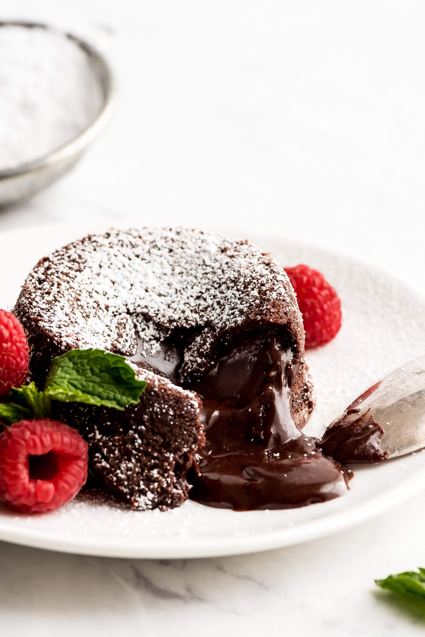 A Molten Lava Cake on a plate, dusted with powdered sugar, chocolate oozing out, and raspberries on the side.