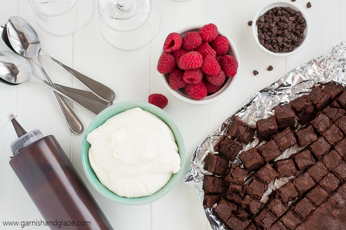 Tablescape of ingredients for Raspberry Brownie Cheesecake Trifles- brownies, raspberries, chocolate chips, chocolate syrup, and cream cheese.