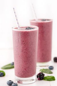 Purple Berry Spinach Smoothie in tall glasses with purple polka dot paper straws and spinach and berries scattered on table.
