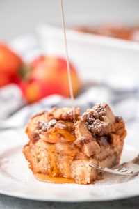 One plate square slice of Overnight Apple French Toast Casserole topped with powdered sugar and maple syrup being drizzled over the top.