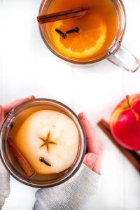 Two glass mugs of slow cooker wassail with an apple slice, orange, slice, cloves, and cinnamon stick floating on top. Hands are wrapped around one of the mugs.