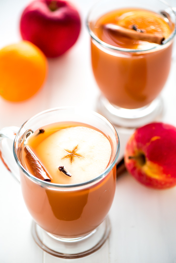 Glass mugs of slow cooker wassail with an apple slice, orange, slice, cloves, and cinnamon stick floating on top.