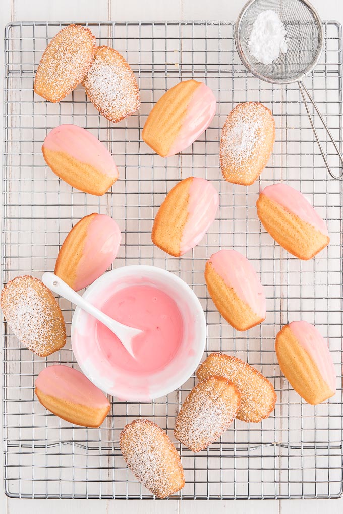 Madeleines on a cooling rack dusted with powdered sugar and half dipped in pink glaze.