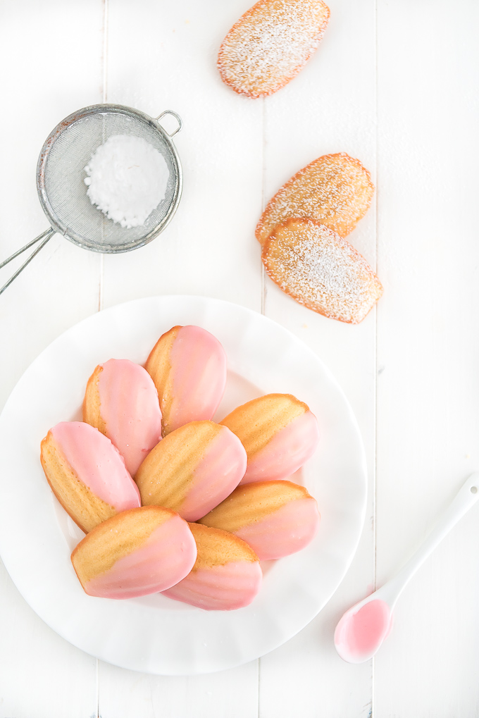 Pink glazed French Madeleines on a plate and dusted ones on the table.