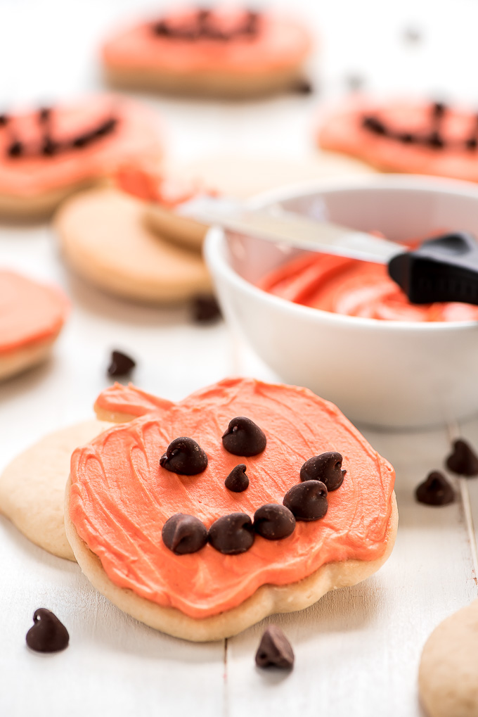 Close up of one Pumpkin Shaped Sugar Cookie frosted with orange butter cream frosting and topped with chocolate chips in the shape of a smiley face.