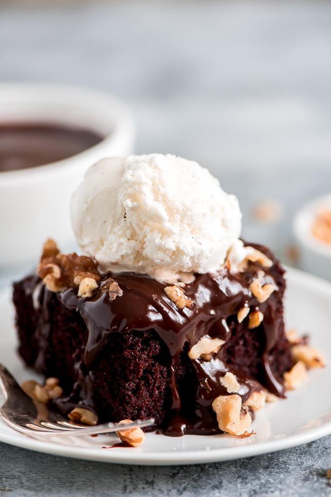A square slice of Chocolate Mayo Cake topped with fudge sauce, walnuts, and a scoop of vanilla ice cream.