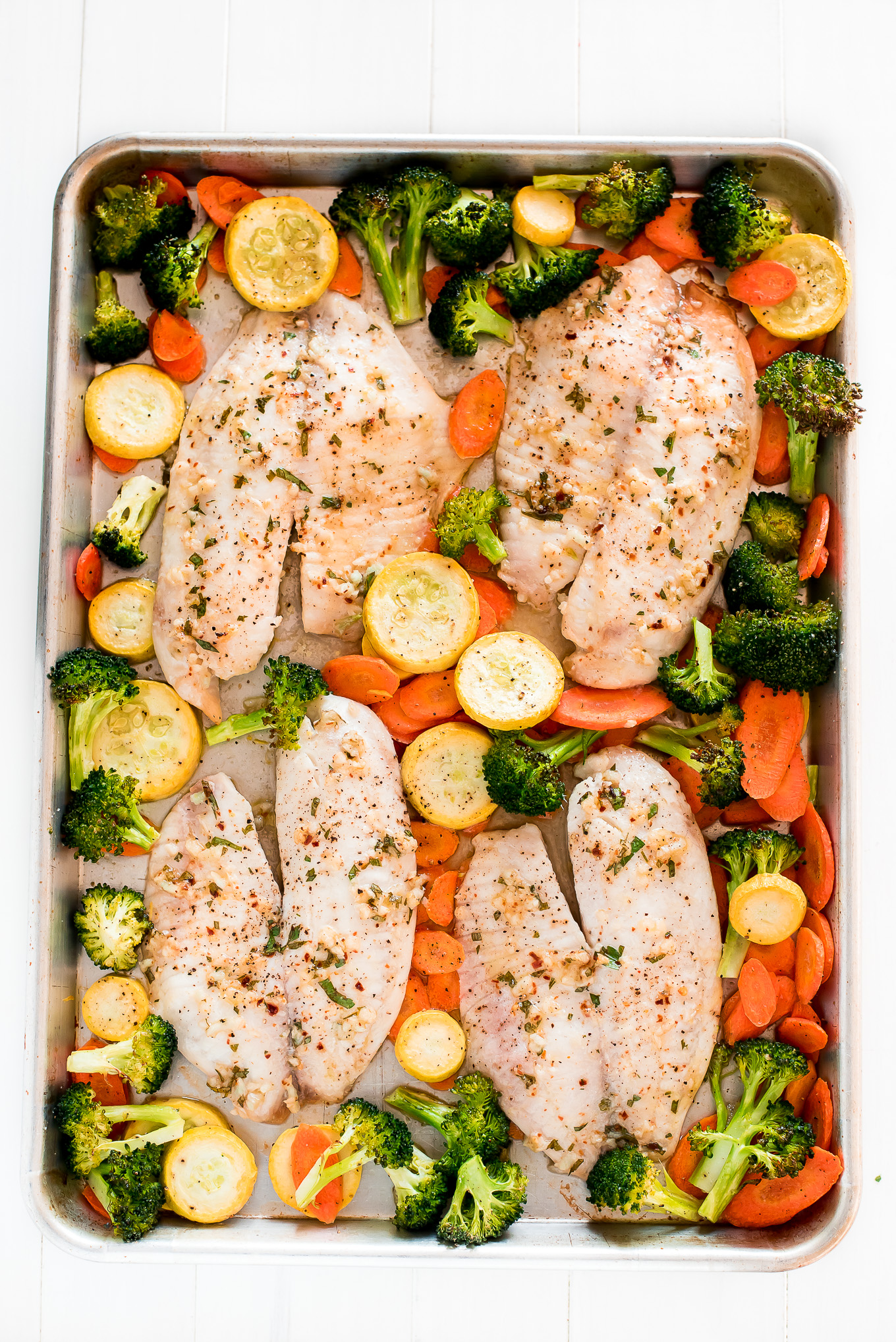 Four baked tilapia fillets and vegetables on a sheet pan.