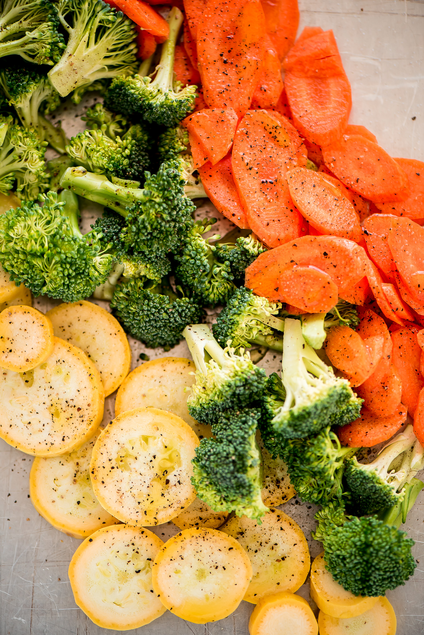 Raw carrot slices, broccoli, and summer squash on a sheet pan, sprinkled with salt and pepper.