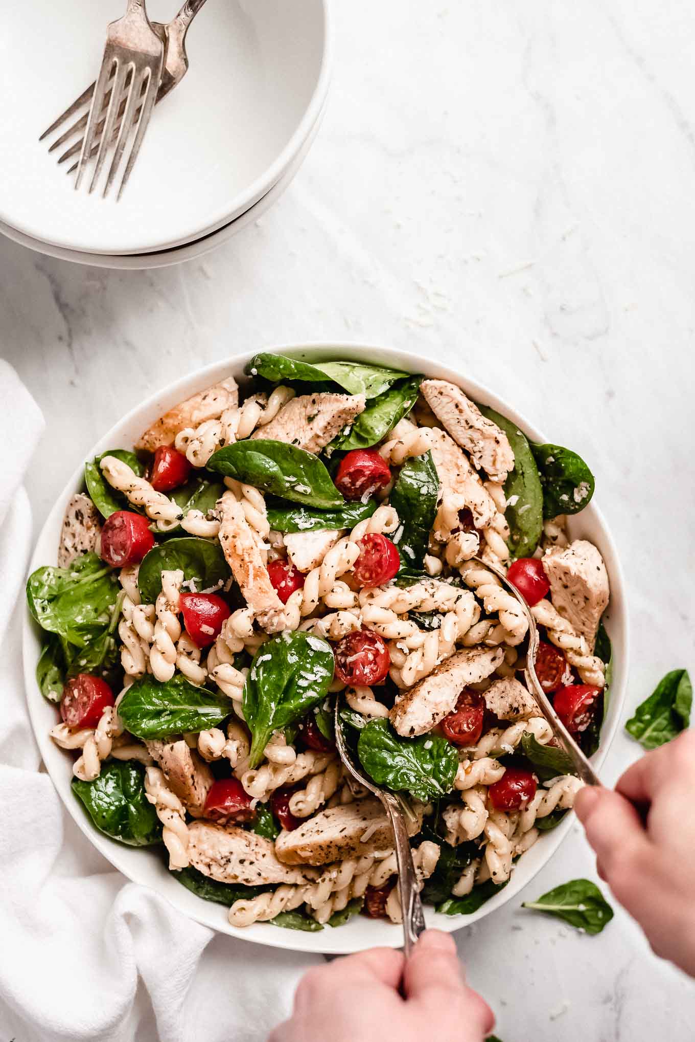  Chicken Pasta Salad in a large bowl. Hands holding two spoons to lift a portion out of the bowl.