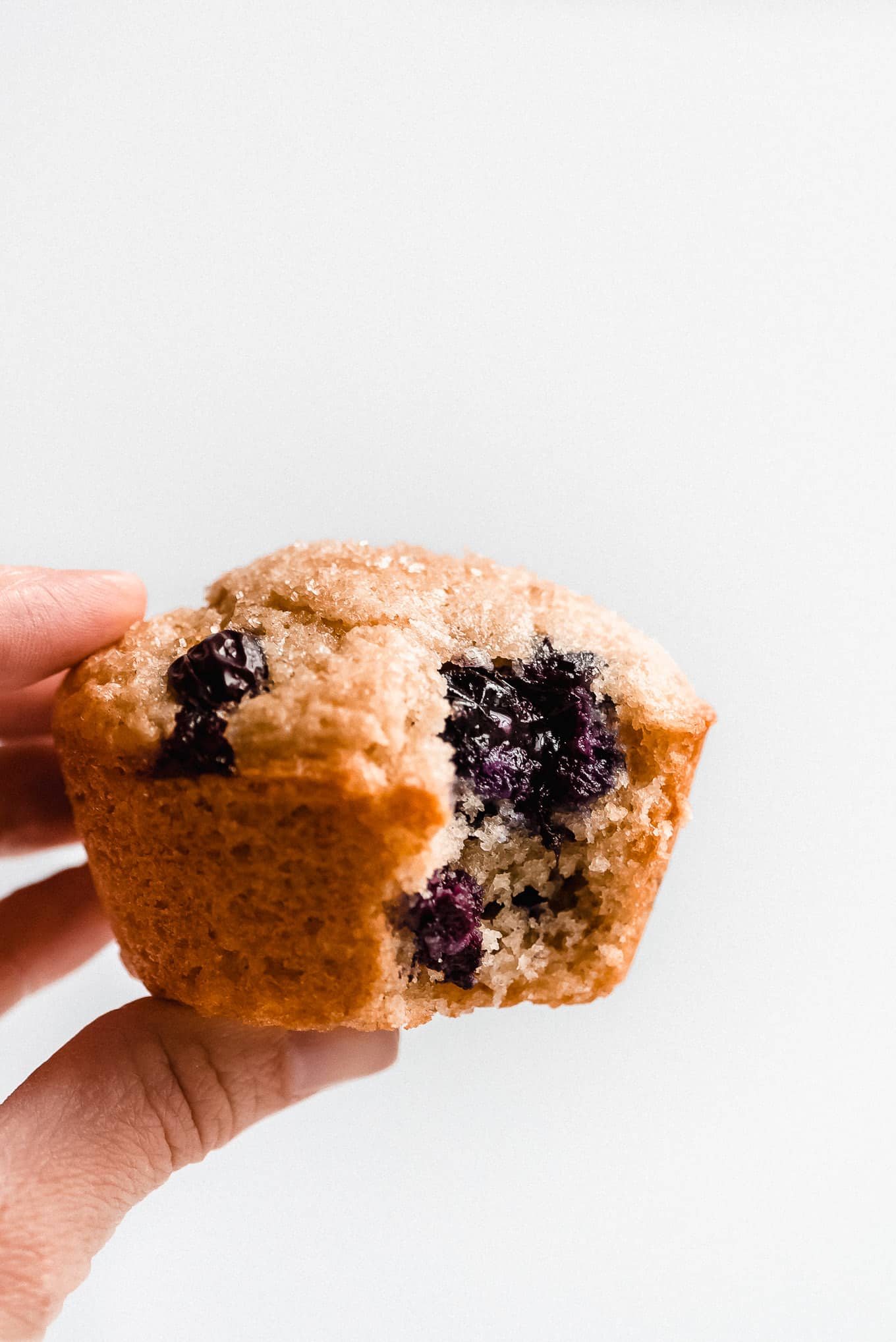 Holding a Greek Yogurt Blueberry Muffin that has a bite taken out, revealing a burst blueberry and the moist tender crumb of the muffin.