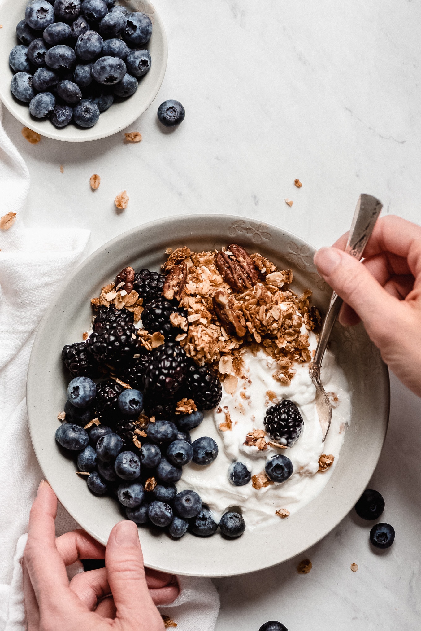 Top view of a bowl of plain Greek yogurt, topped with blackberries, blueberries, and granola with a hand scooping out a bite with a spoon.