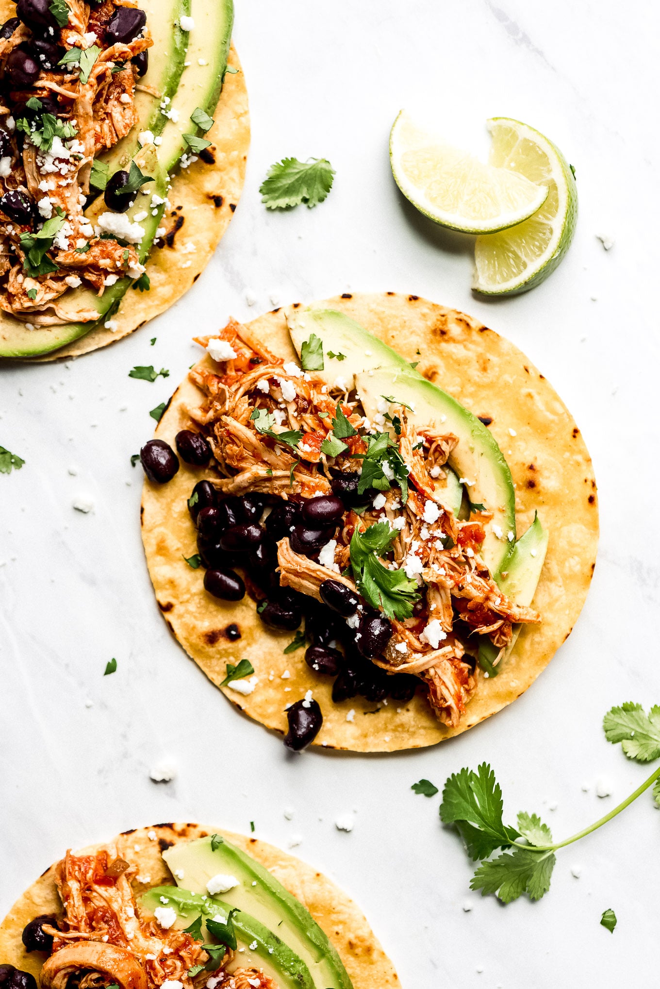 Corn tortillas on a table topped with shredded Salsa Chicken, black beans, avocado slices, cilantro, and Queso Fresco.