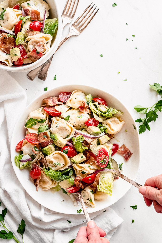 BLT Tortellini Salad in a large bowl and a person using two spoons to scoop some out.