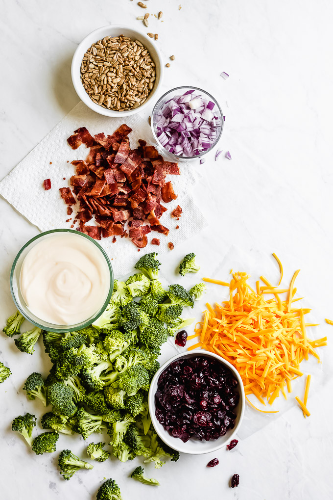 Ingredients for making broccoli salad with bacon on a marble surface- sunflower seeds, red onion, chopped bacon, creamy dressing, broccoli florets, cheddar cheese, and dried cranberries.