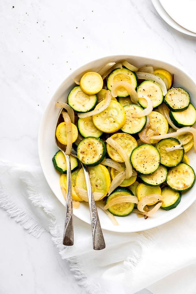 Sautéed Zucchini and Summer Squash in a serving bowl with a cloth napkin at the side and two spoons in the bowl.