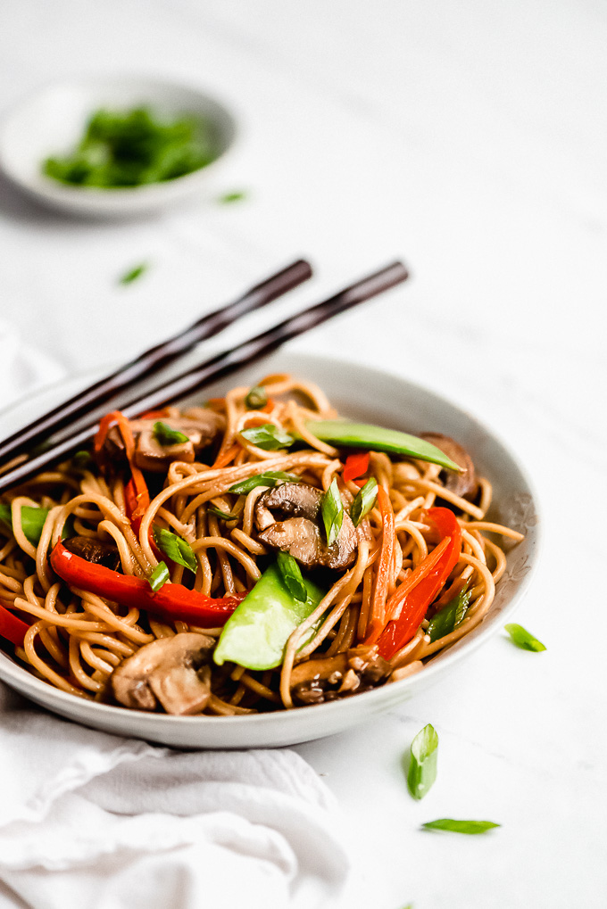 A bowl of Vegetable Lo Mein with chop sticks resting on the edge of the bowl and green onions scattered on the table.