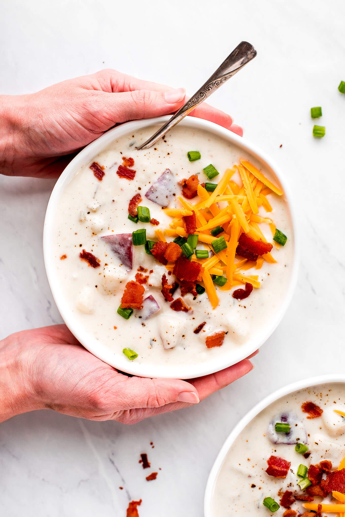 Two hands on the sides of a bowl of potato soup topped with cheese, bacon, and green onion.