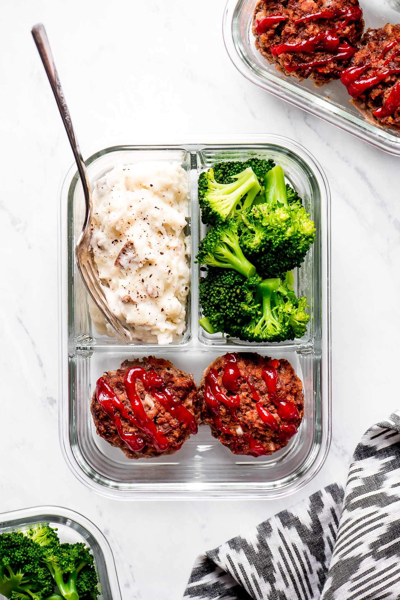 A glass meal prep container with two mini meatloaf, mashed potatoes, and broccoli.