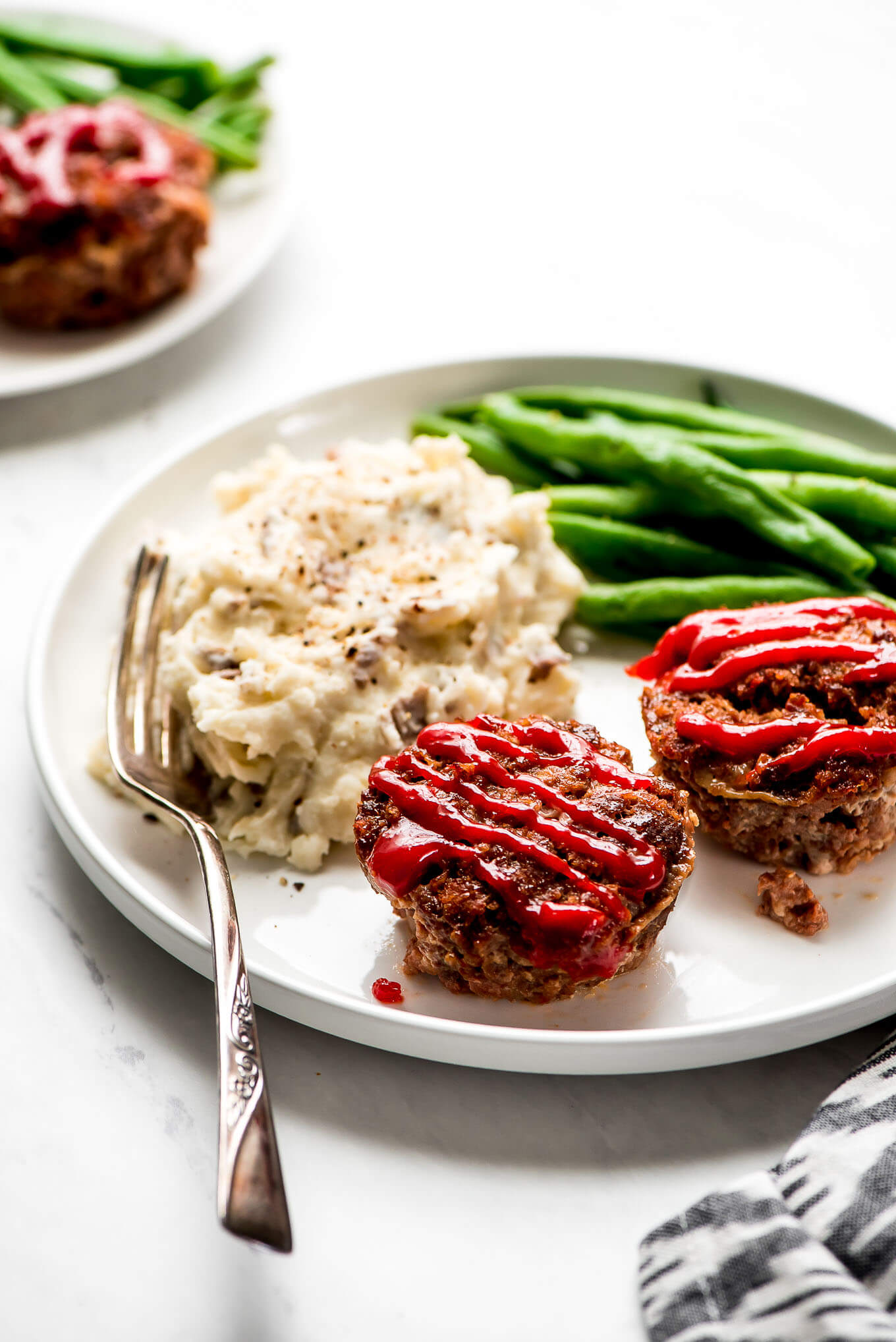 A dinner plate with focus on Mini Meatloaf and mashed potatoes and green beans in the background with a fork on the side.