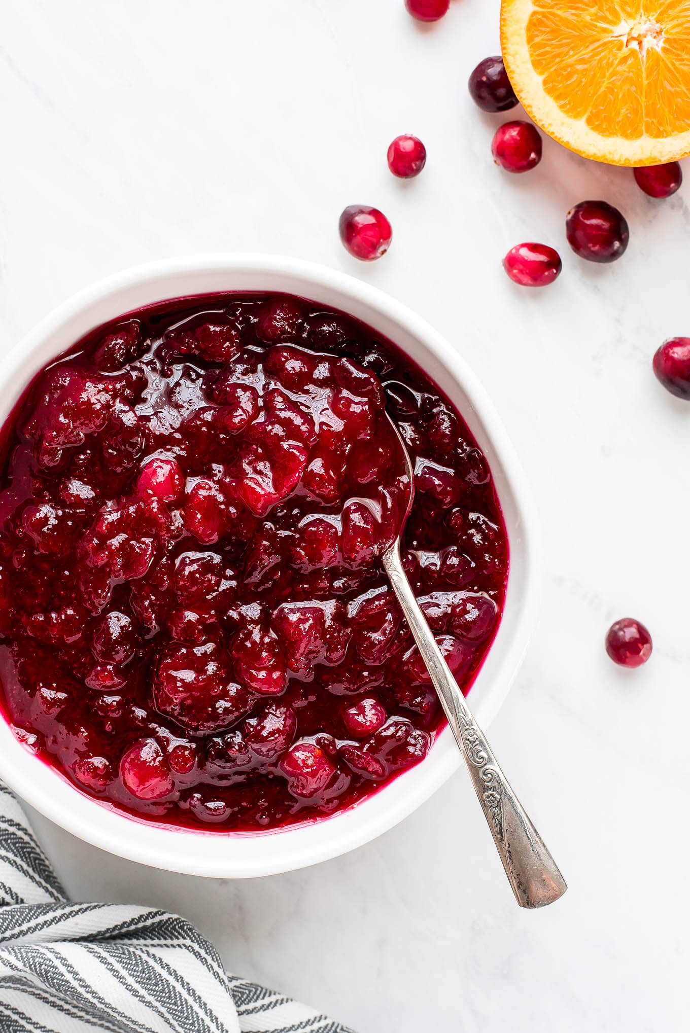 Cranberry sauce in a bowl with a spoon and a half orange and cranberries to the side.