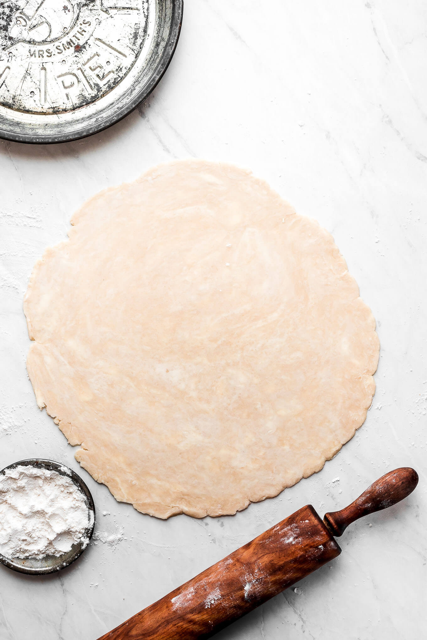 Roll out dough for making homemade pie crust and a rolling pin, pie tin, and dish of flour to the side.