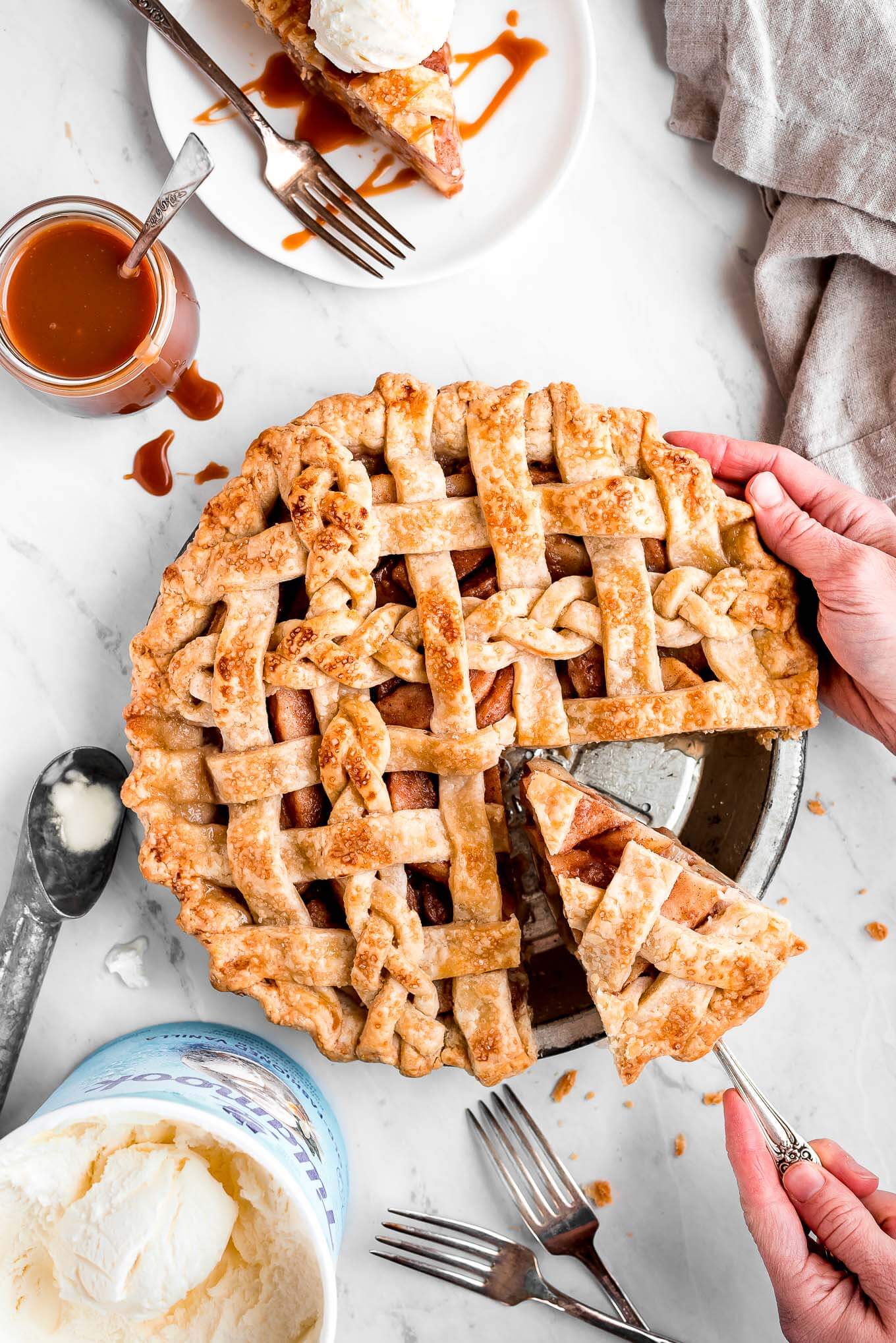 Pear Pie with a carton of vanilla ice cream, ice cream scoop, jar of caramel sauce, and a slice of pie surrounding it with a hand lifting out a slice of pie with a pie server.