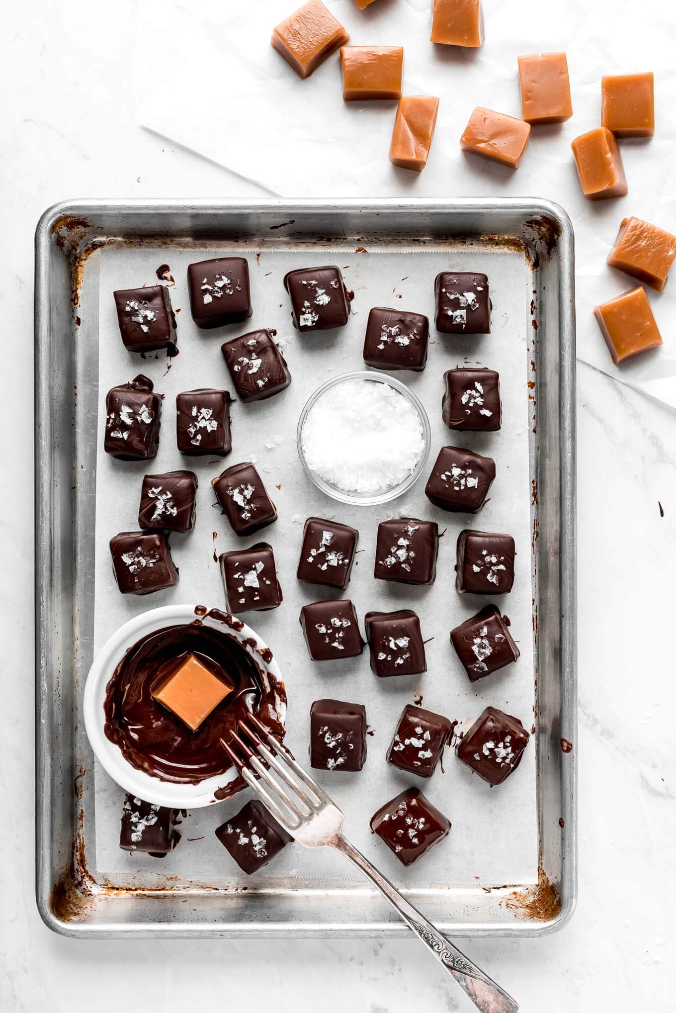 A baking sheet full of chocolate covered caramels, a dish of melted chocolate, and a small bowl of sea salt.
