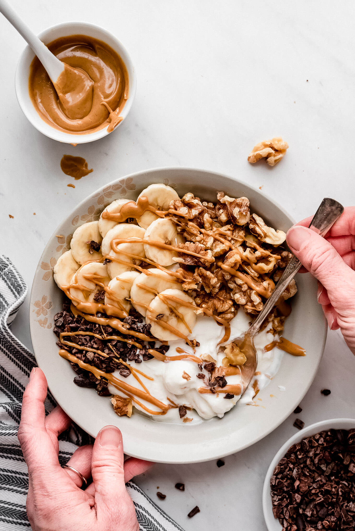Hands holding a bowl and lifting a spoonful of yogurt from a Greek yogurt bowl topped with bananas, chocolate, and nuts.