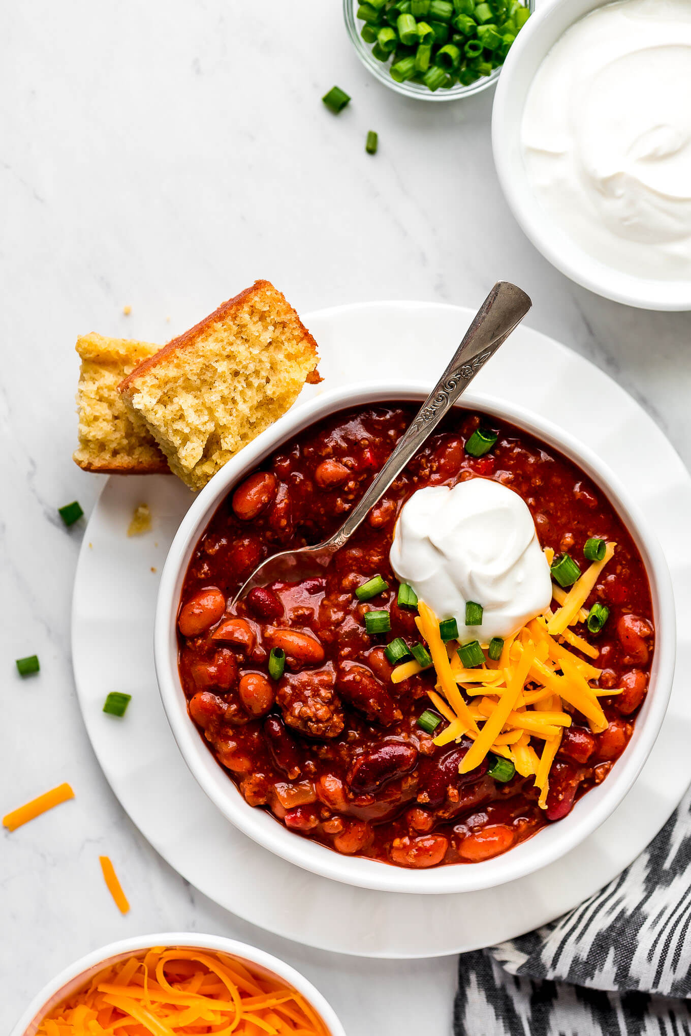 Instant Pot Chili in a bowl on a plate and topped with cheddar cheese, sour cream, and green onions. Corn bread and bowls of toppings to the side.
