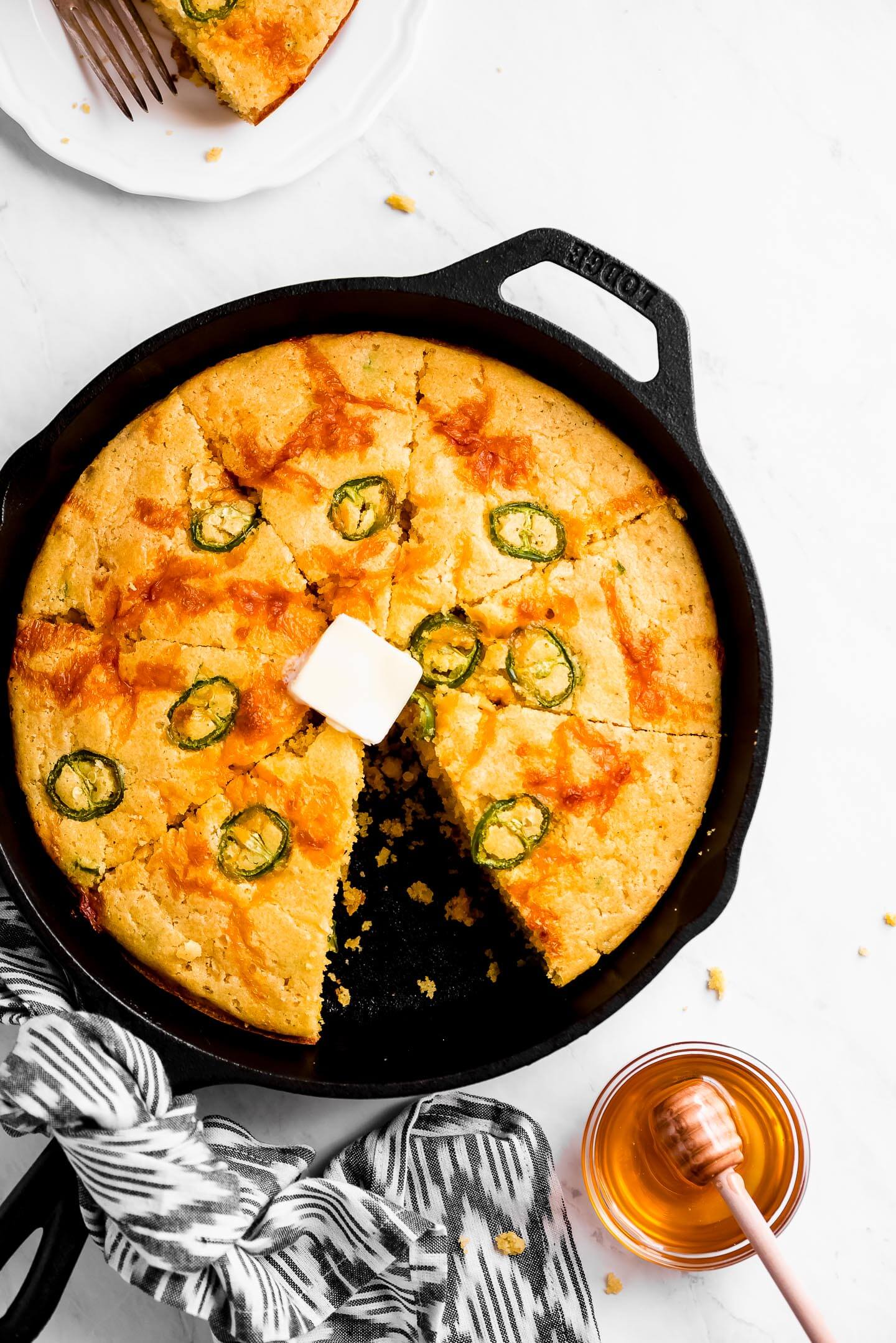Jalapeno Cheddar Cornbread in a skillet sliced into triangles and a piece taken out and on a plate to the side.