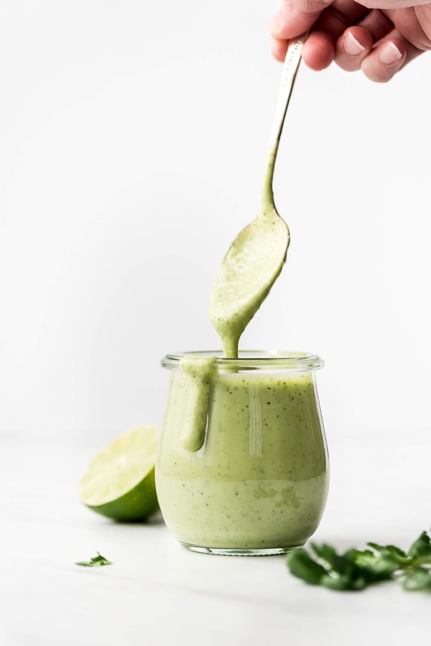 A hand lifting a spoon out of a jar of green dressing that has some dripping down the front of the jar.