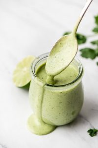 A jar of Cilantro Lime Dressing with a spooning being lifted out and dressing dripping back into the jar.