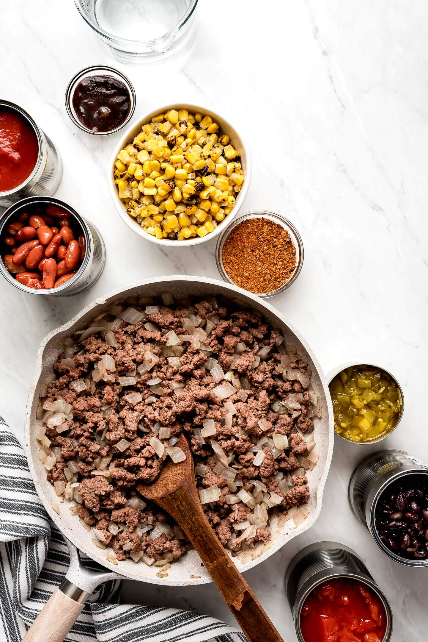 A skillet of browned ground beef and onions, bowl of frozen corn, seasoning, cans of beans, diced green chilies, tomatoes, and tomato sauce.