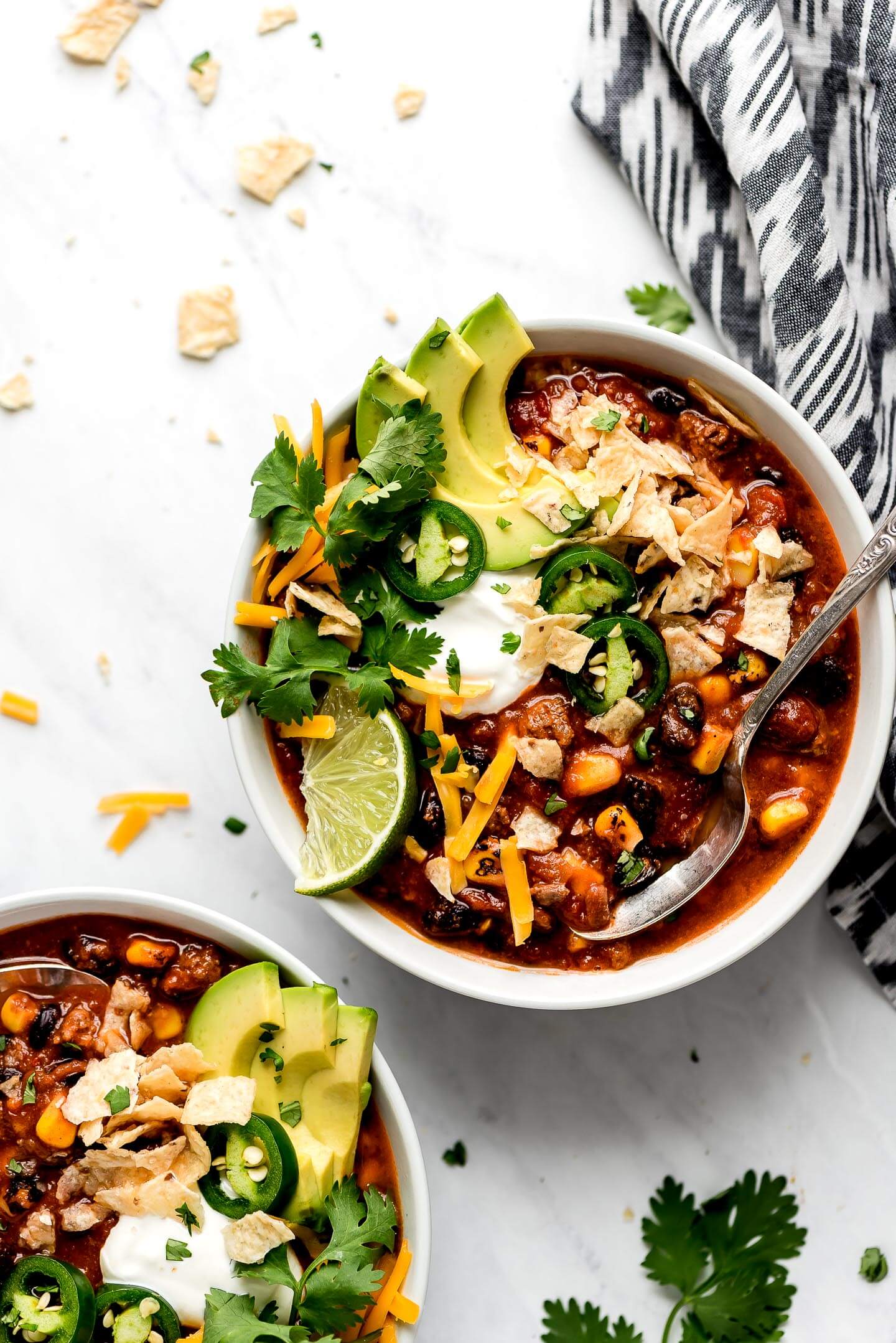Two bowls of Slow Cooker Taco Soup topped with avocado slices, cilantro, sliced jalapenos,chips, and a a lime wedge and cloth napkin to the side.