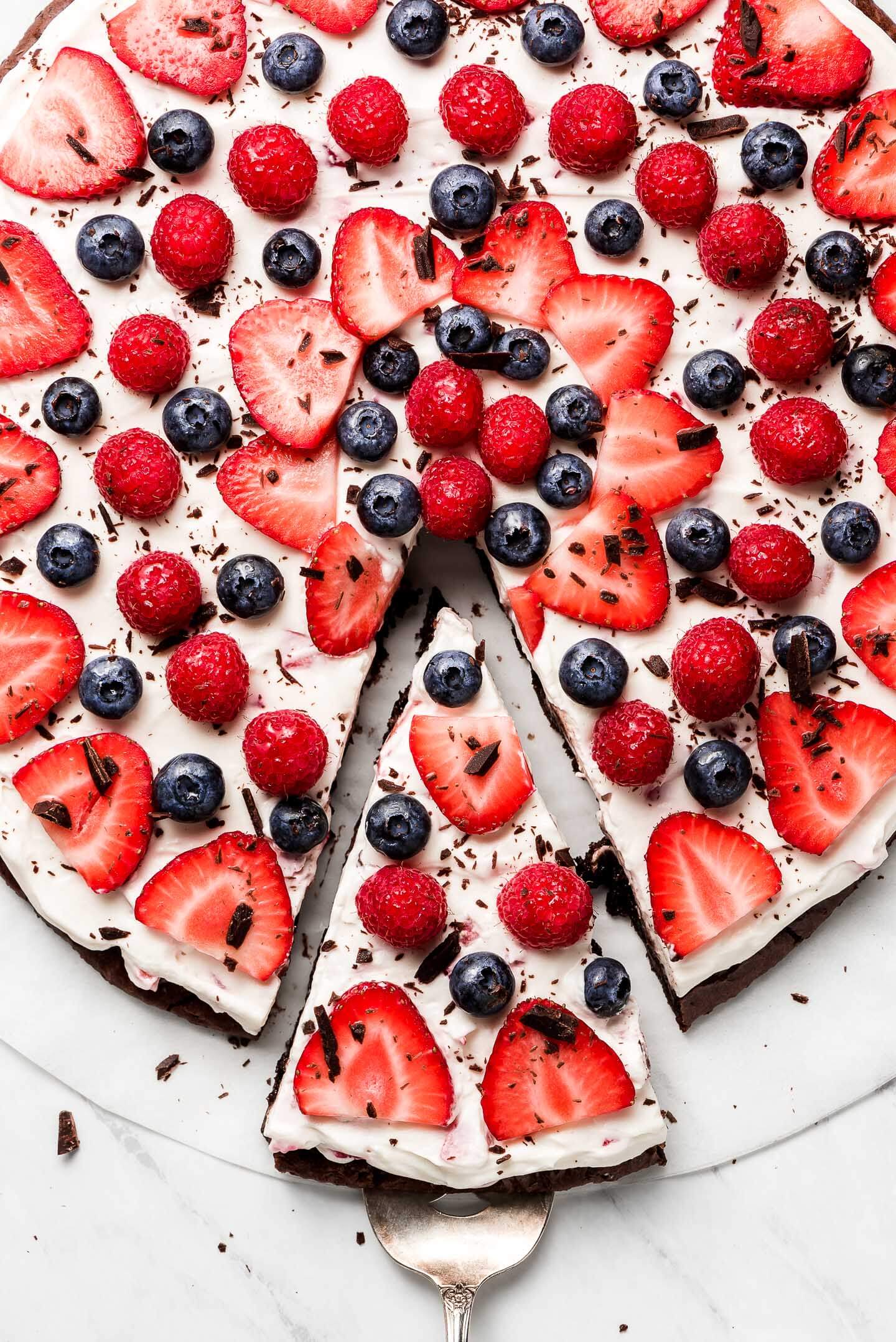 Berry Brownie Pizza decorated with strawberries, raspberries, and blueberries with a slice cut out and being pulled away.