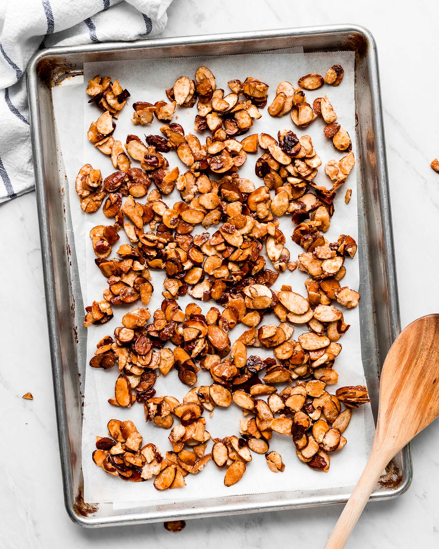 Caramelized nuts on a baking sheet with a wooden spoon resting on the corner and kitchen towel at the side.