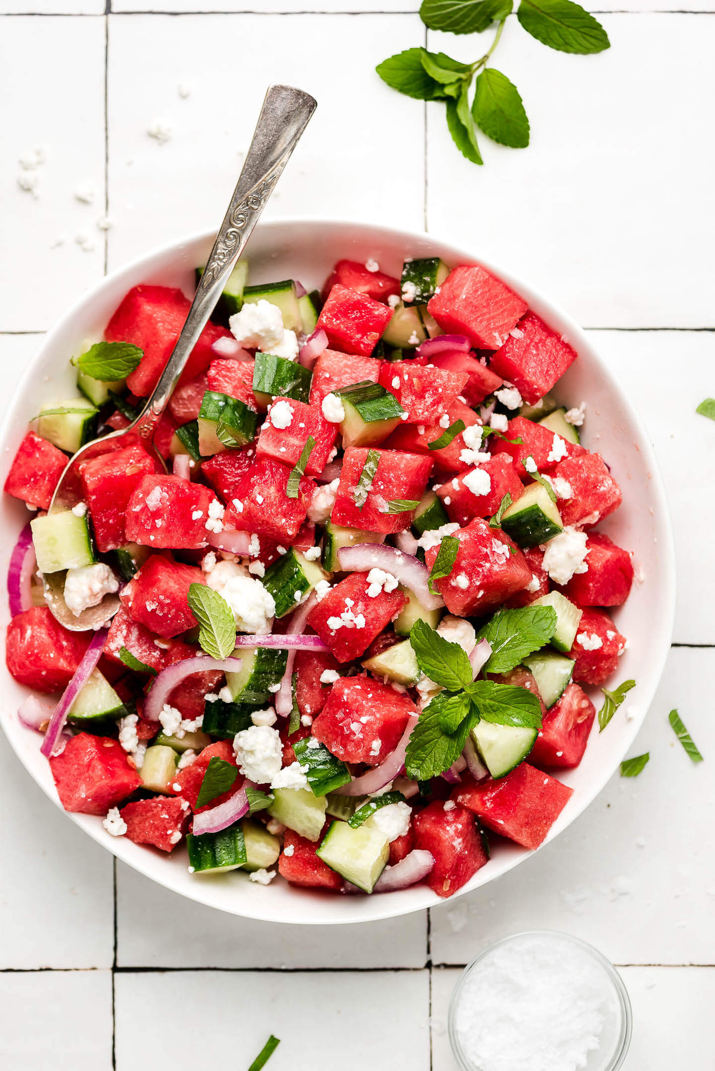 A large white bowl of Watermelon Feta Salad with a small bowl of salt to the side and a sprig of mint on the table.