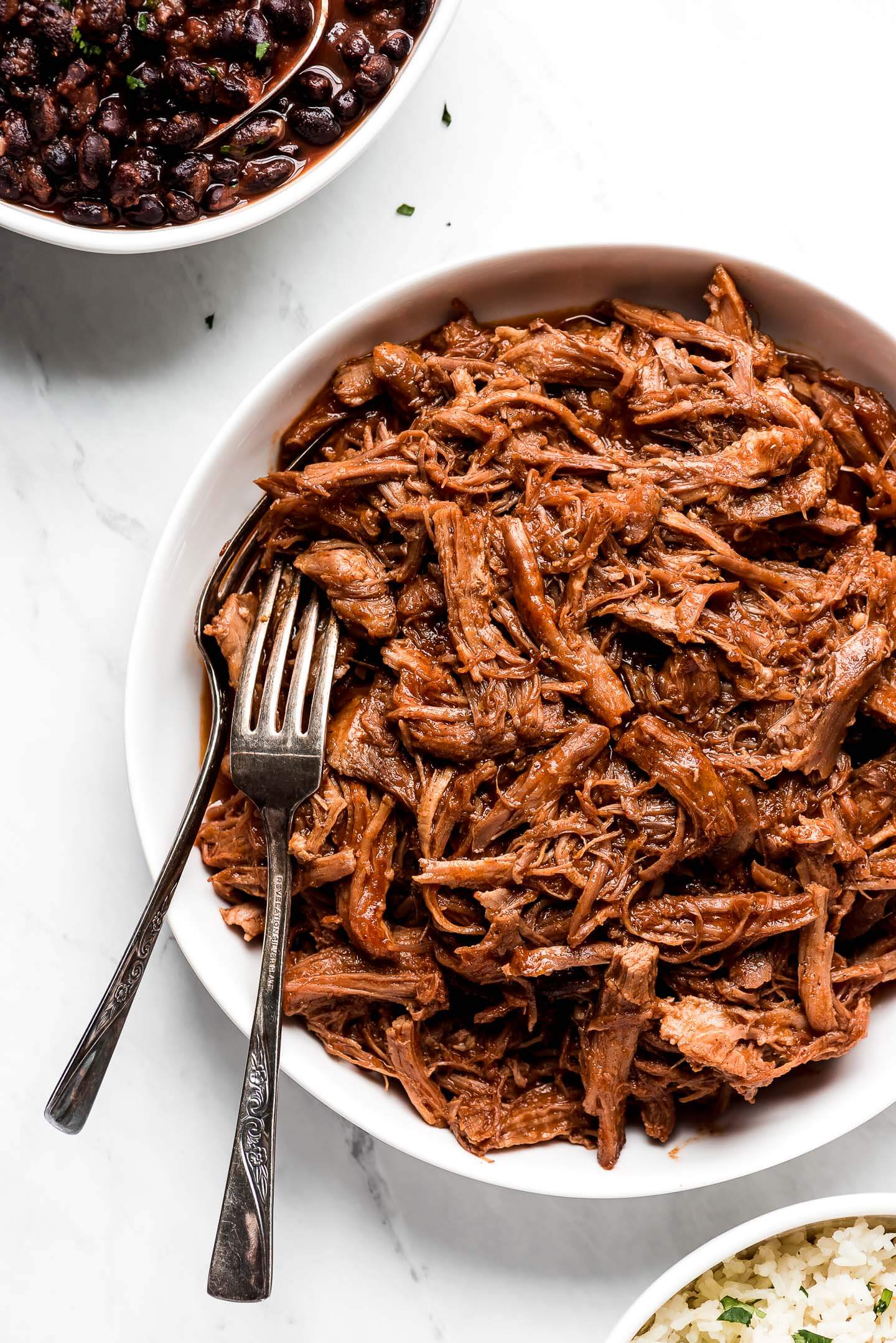Sweet pulled pork in a bowl with two forks for serving and bowls of black beans and cilantro lime rice to the sides.