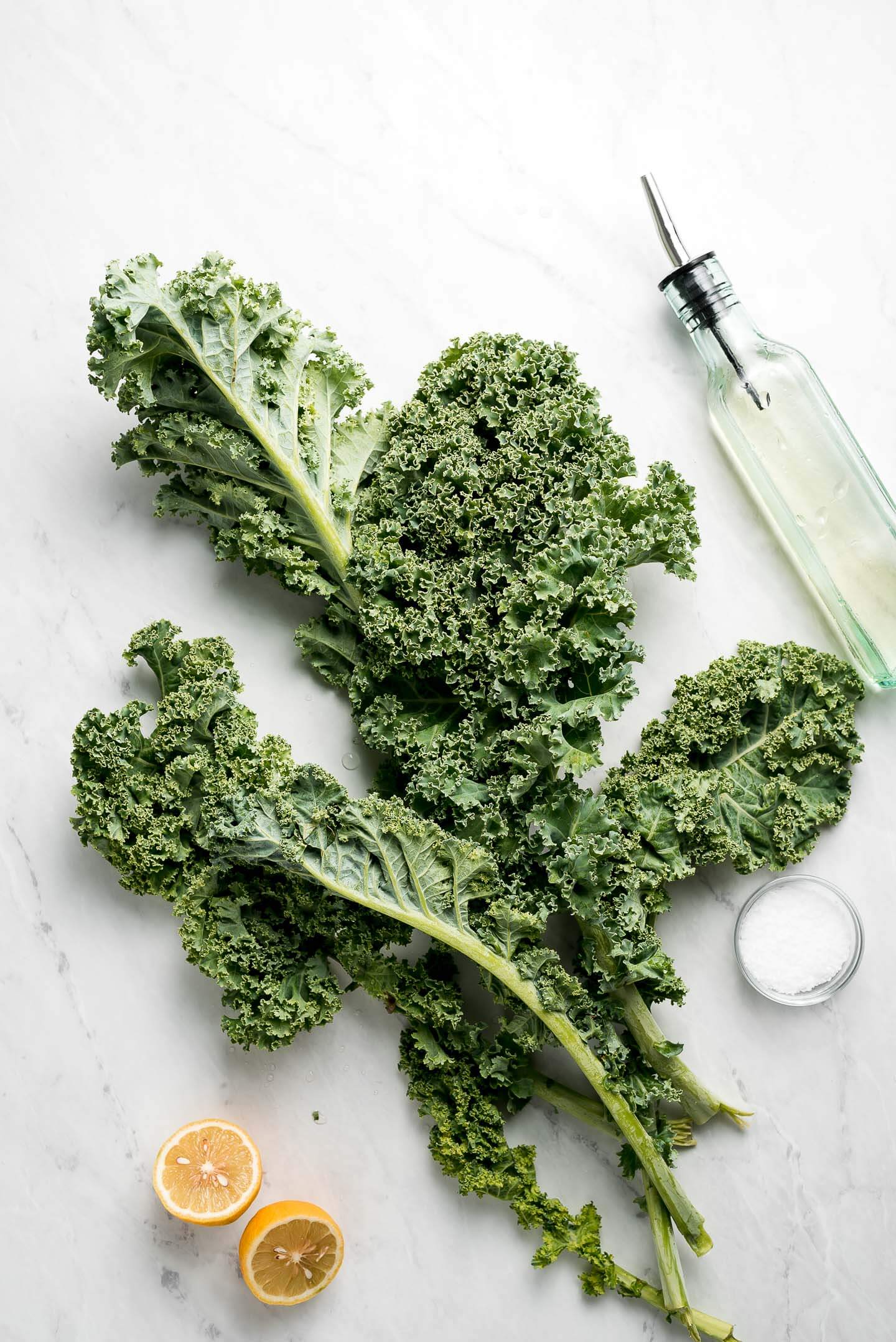 Leaves of raw kale on a marble surface with a bottle of oil, lemon halves, and a small bowl of kosher salt.