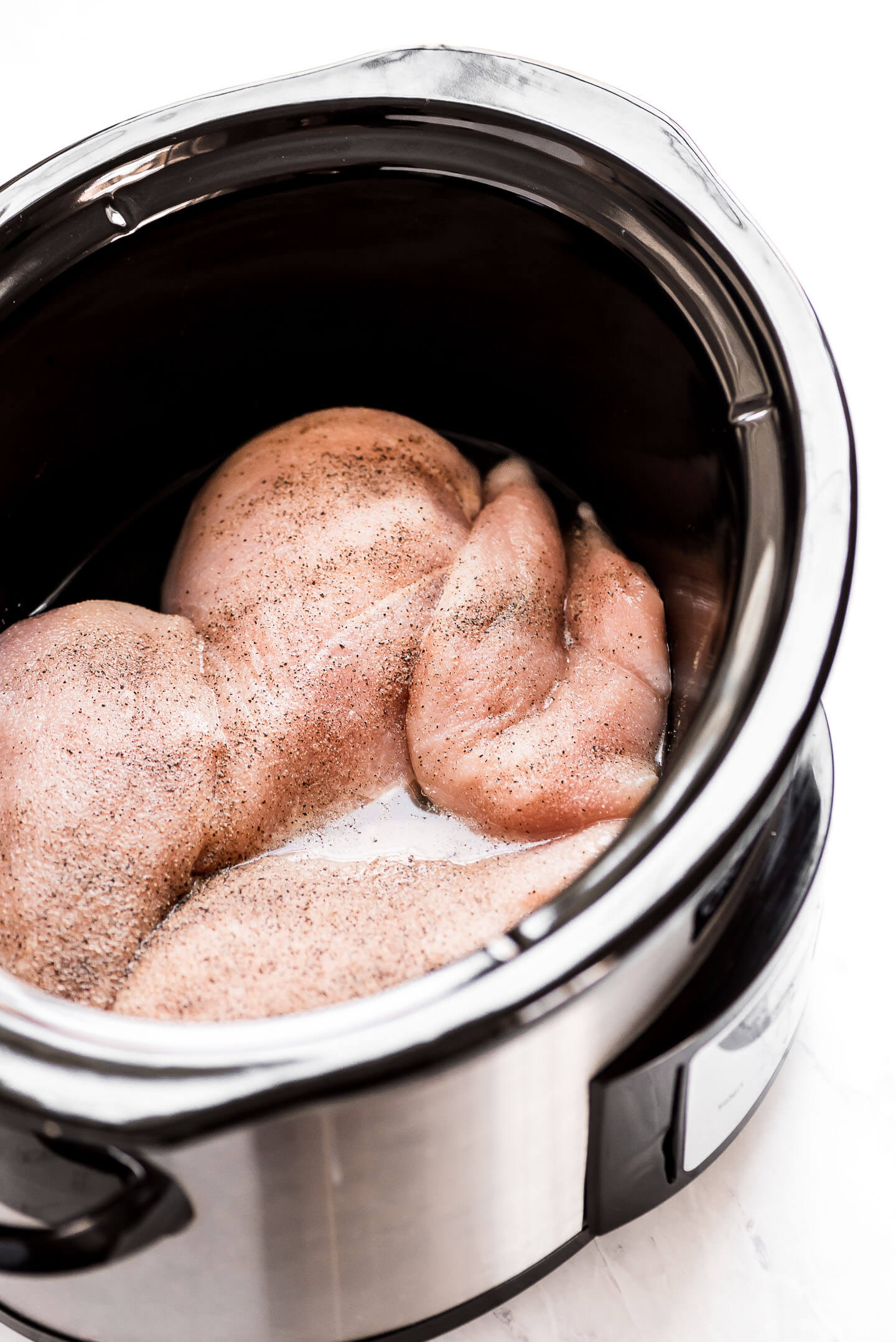 Raw boneless skinless chicken breasts in the bottom of a slow cooker seasoned with spices.