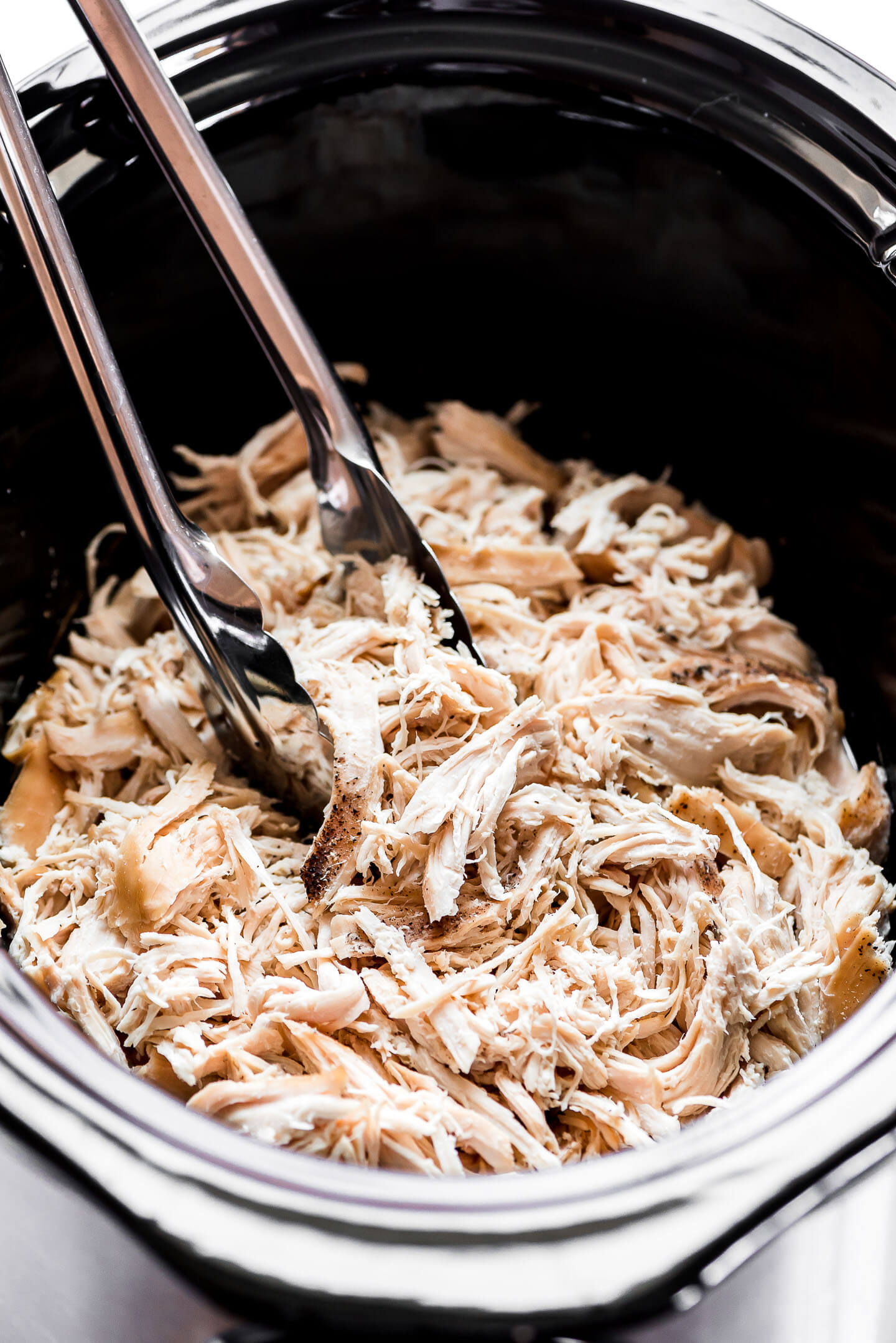 Shredded chicken in a Crock-Pot with tongs about to lift some out.