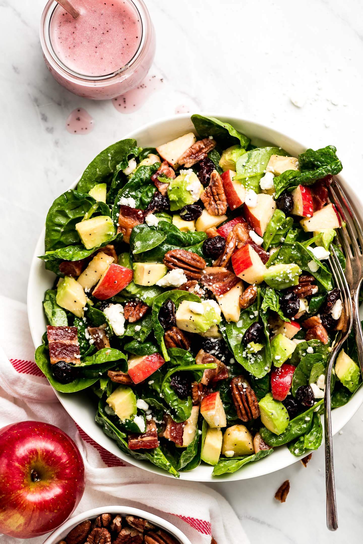 Apple & Bacon Spinach Salad tossed in a poppy seed dressing in a large bowl with an apple and a jar of pink poppy seed dressing to the side.