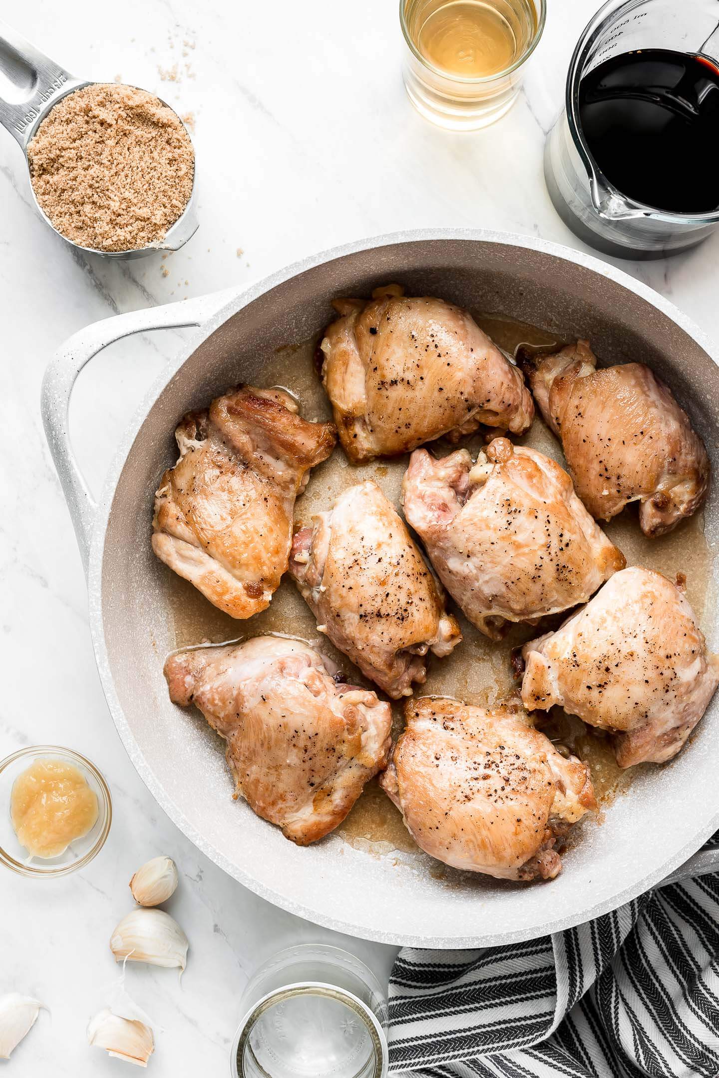 Browned chicken thighs in a skillet seasoned with salt and pepper with various ingredients (brown sugar, soy sauce, vinegar, garlic, and ginger) surrounding.