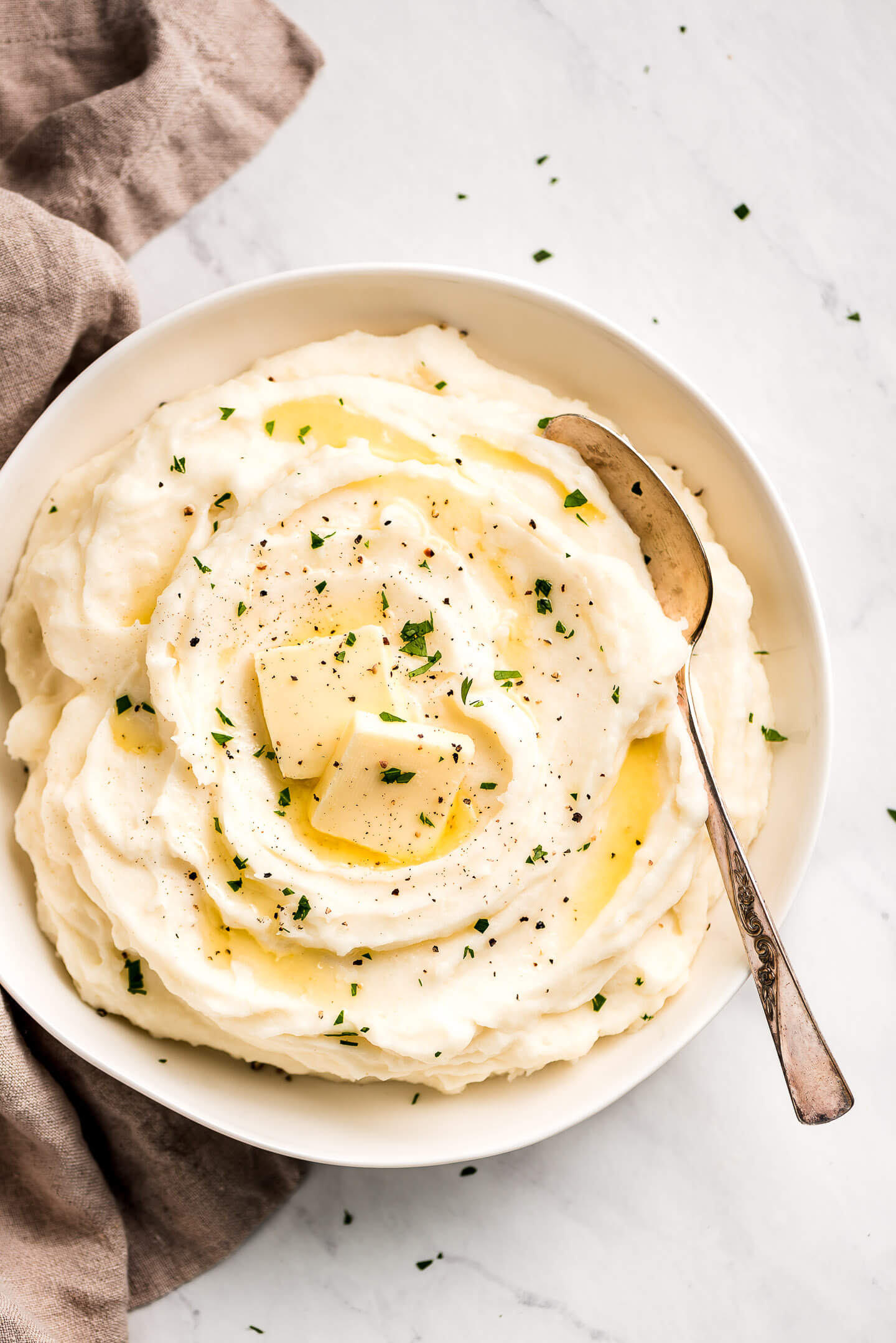 The best Mashed Potatoes garnished with pats of butter, pepper, and parsley in a large white serving bowl.