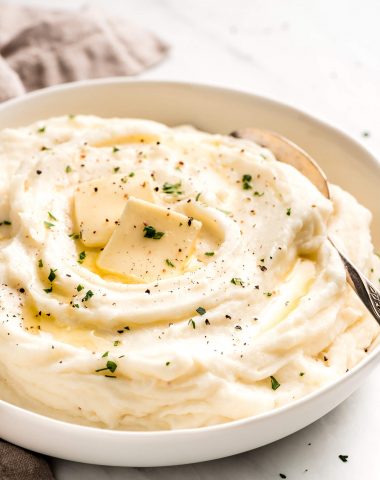 Creamy Mashed Potatoes in a large white serving bowl and topped with pats of butter, parsley, and pepper.