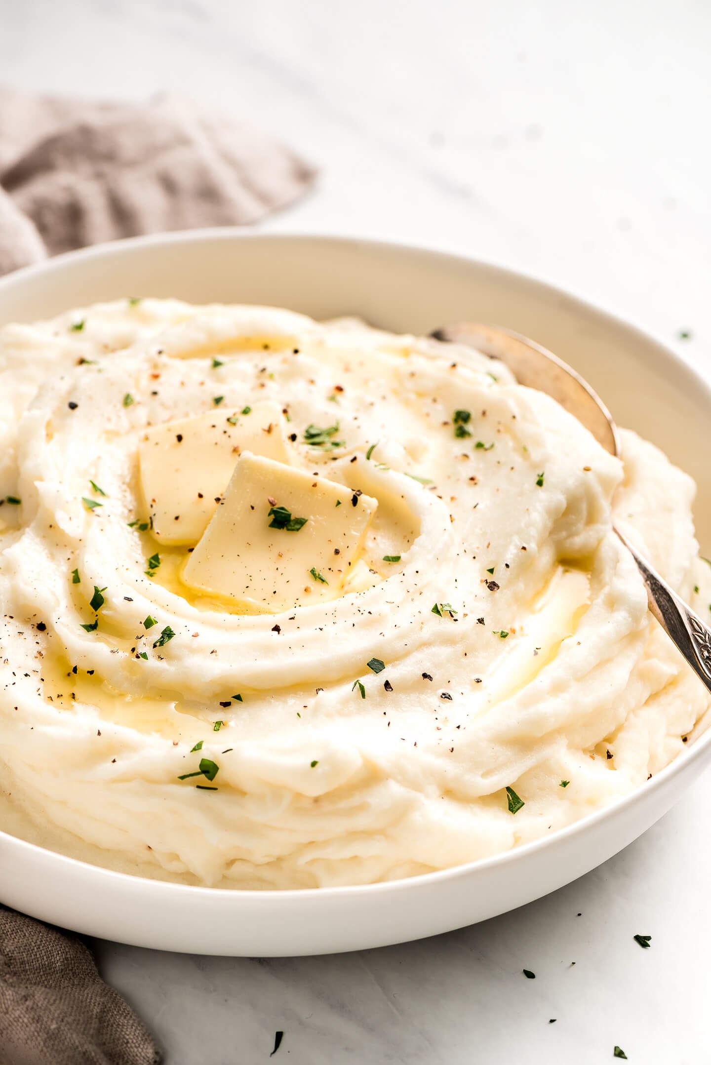 Creamy Mashed Potatoes in a large white serving bowl and topped with pats of butter, parsley, and pepper.