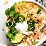 White Chicken Chili in a bowl topped with avocado slices, cilantro, jalapeno, chips, cheese, sour cream, and a lime.