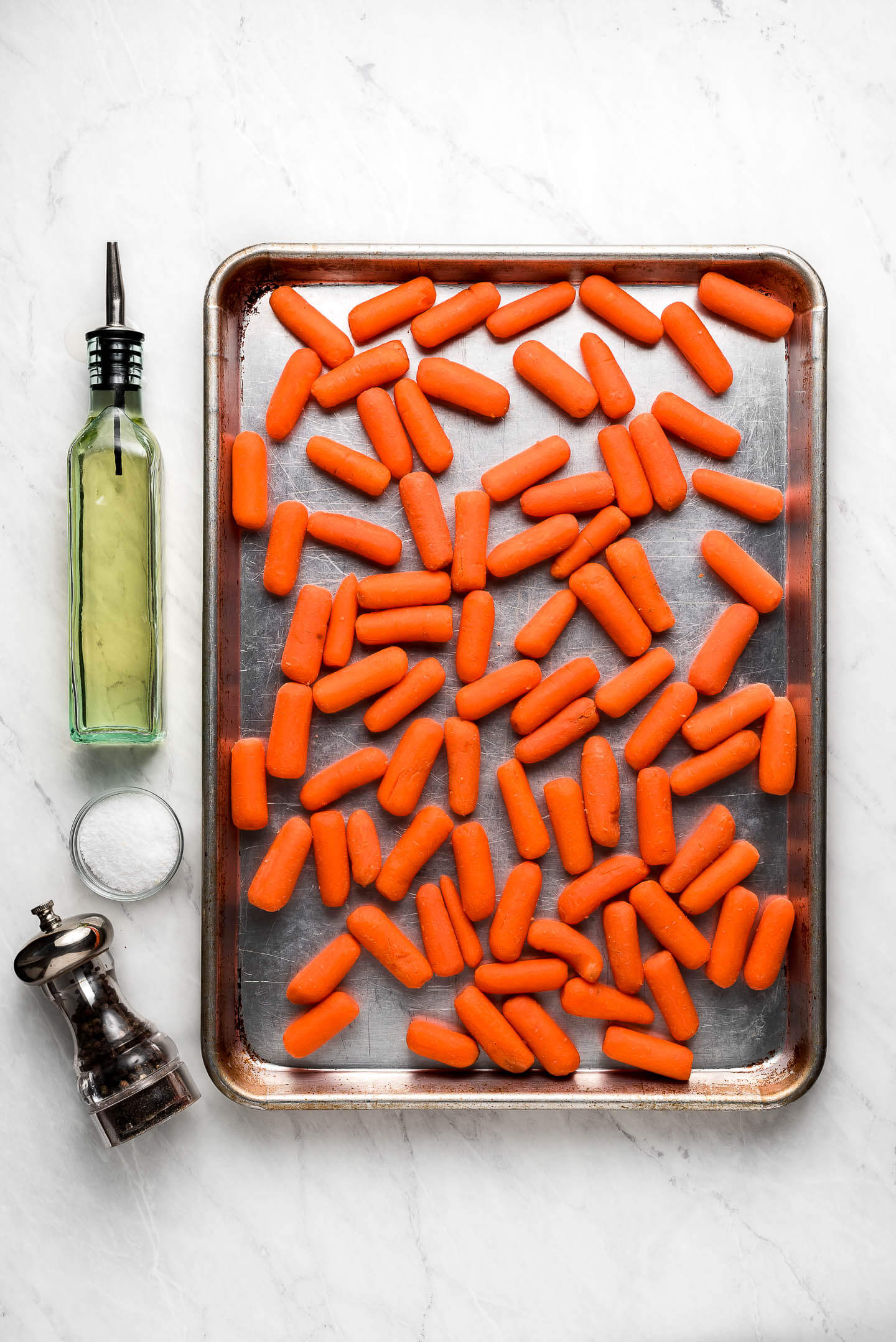 Baby carrots on a baking sheet, a bottle of oil, bowl of salt, and pepper grinder.